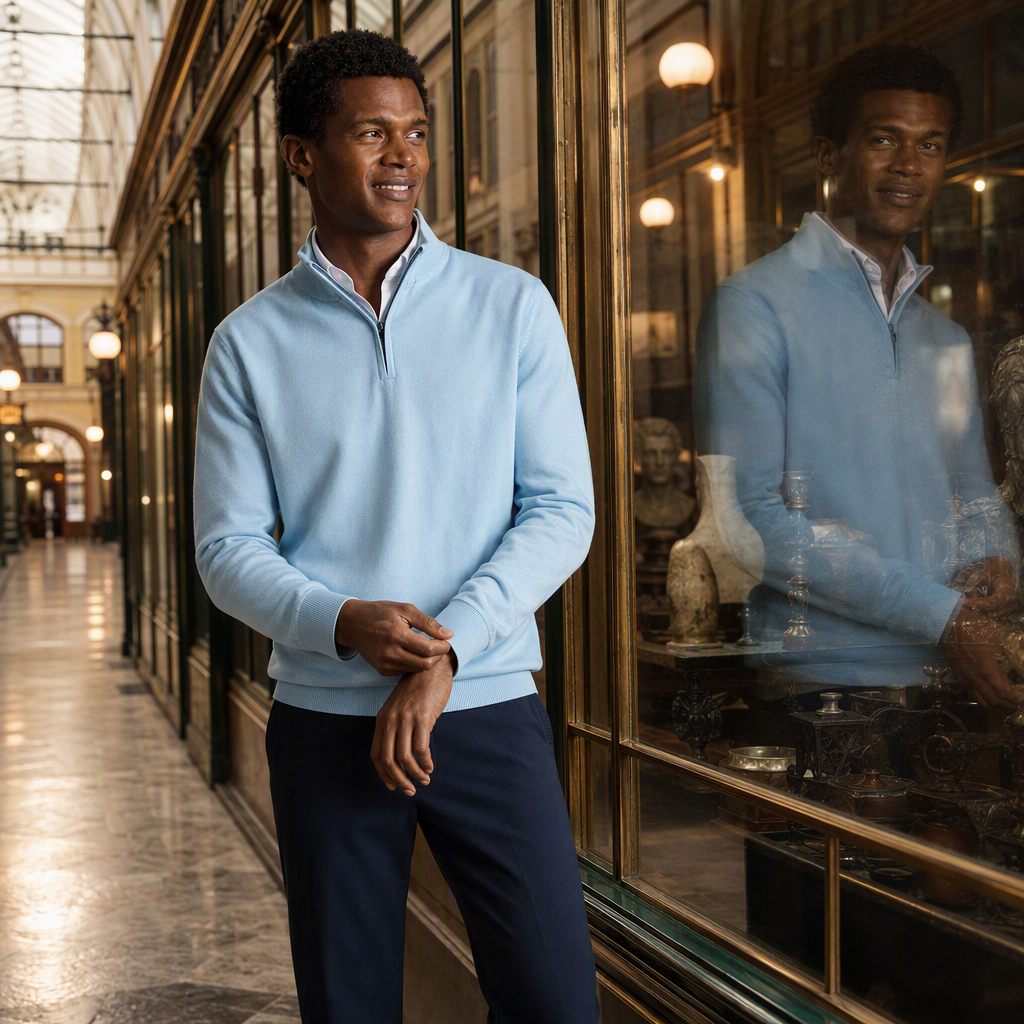 AI-generated product photograph for Charles Tyrwhitt: a man in a pale blue quarter-zip jumper over a white collar and navy trousers smiles beside a shop window in a lamp-lit arcade, his reflection in the glass