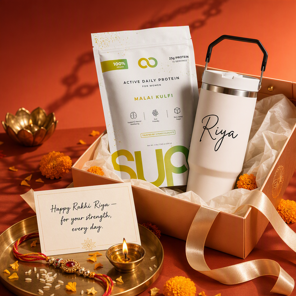 AI product photography for SUPR: a gift box holding a pouch of Malai Kulfi flavour Active Daily Protein for Women and a white tumbler printed with the name Riya, beside a brass plate with a rakhi, a diya and a handwritten card reading "Happy Rakhi Riya, for your strength, every day"